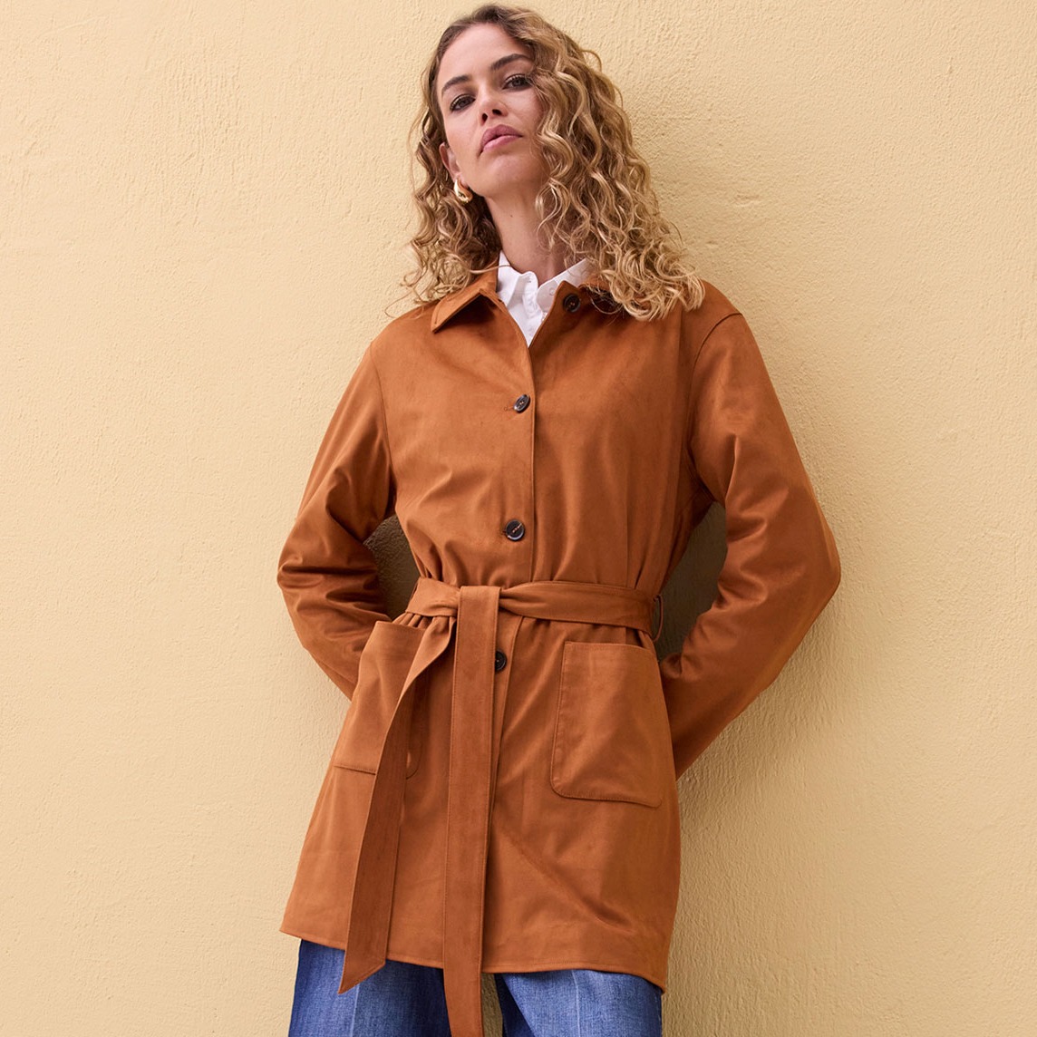 Rostfarbener Mantel mit Gürtel, kombiniert mit weißem Hemd und blauen Jeans.